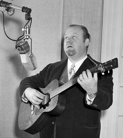 Photograph of Burl Ives singing and playing guitar in a recording studio.